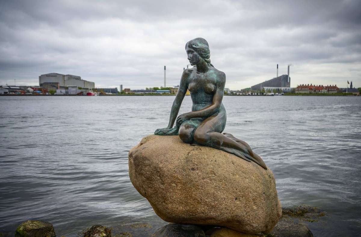 Die kleine Meerjungfrau, Wahrzeichen von Kopenhagen, sitzt auf einem Felsen am Hafen. Im Hintergrund sieht man rechts eines der spektakulärsten Gebäude der dänischen Hauptstadt: das Müllheizkraftwerk Amager Bakke. Auf dem Dach beherbergt es insgesamt 450 Meter an Skipisten (CopenHill). Foto: imago images//Valentin Wolf